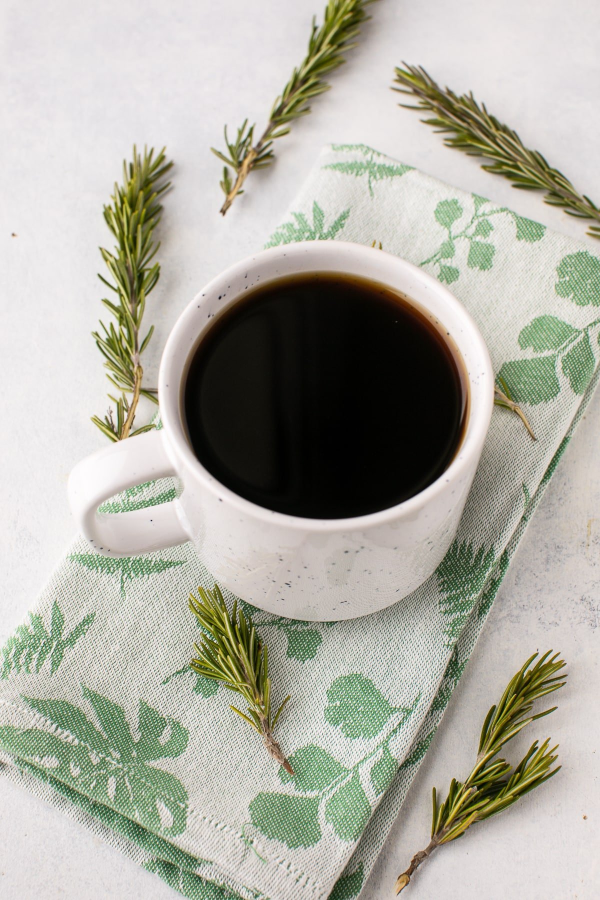 white mug with coffee and sprigs of rosemary on a table.