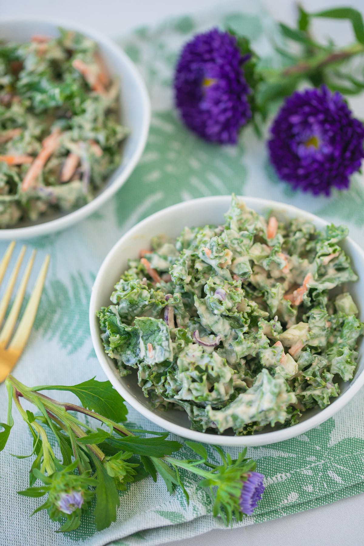 bowl of creamy kale salad on a green floral napkin with fresh purple flowers.