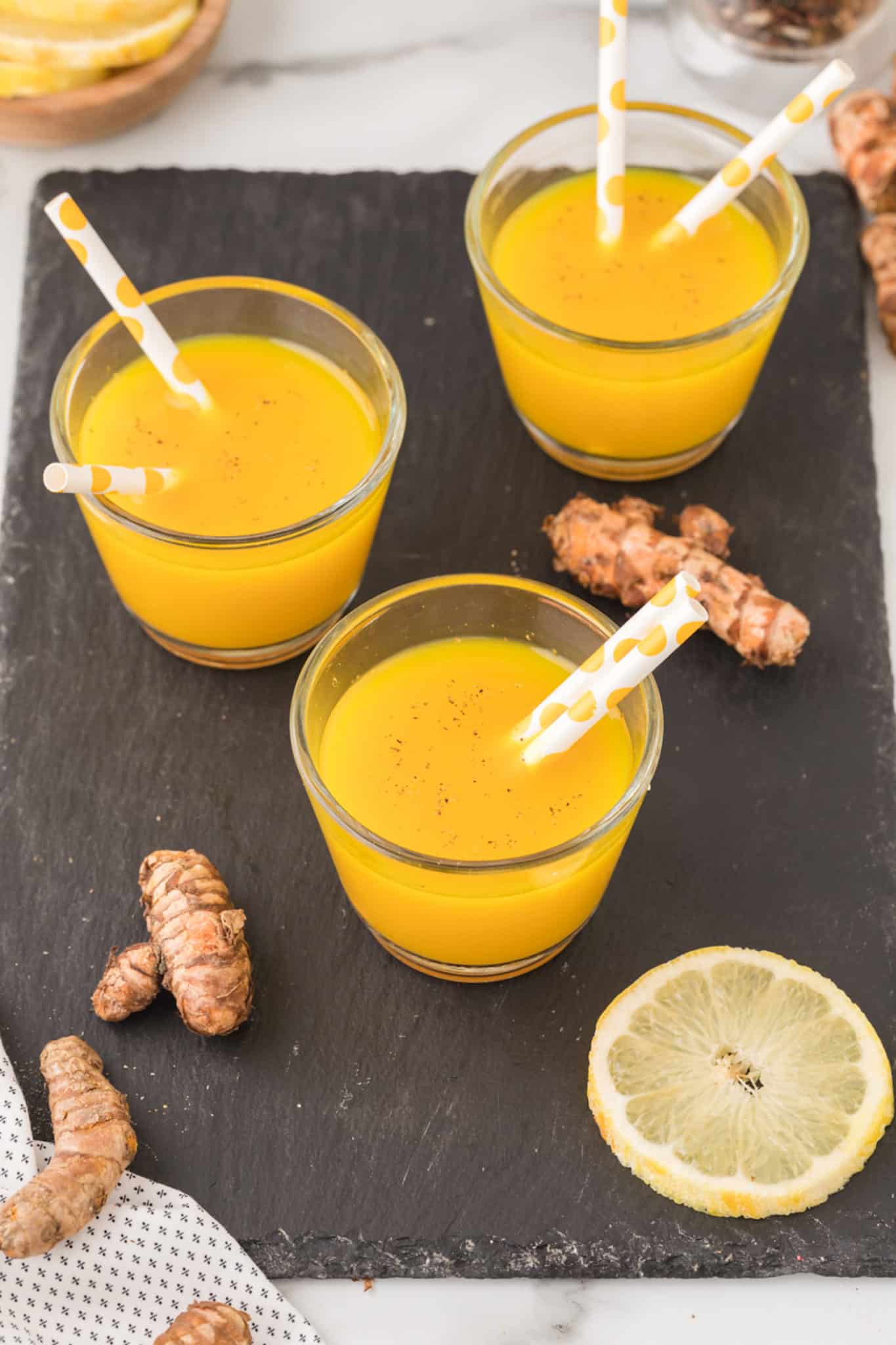 three glasses of yellow turmeric shot with straws on a table.