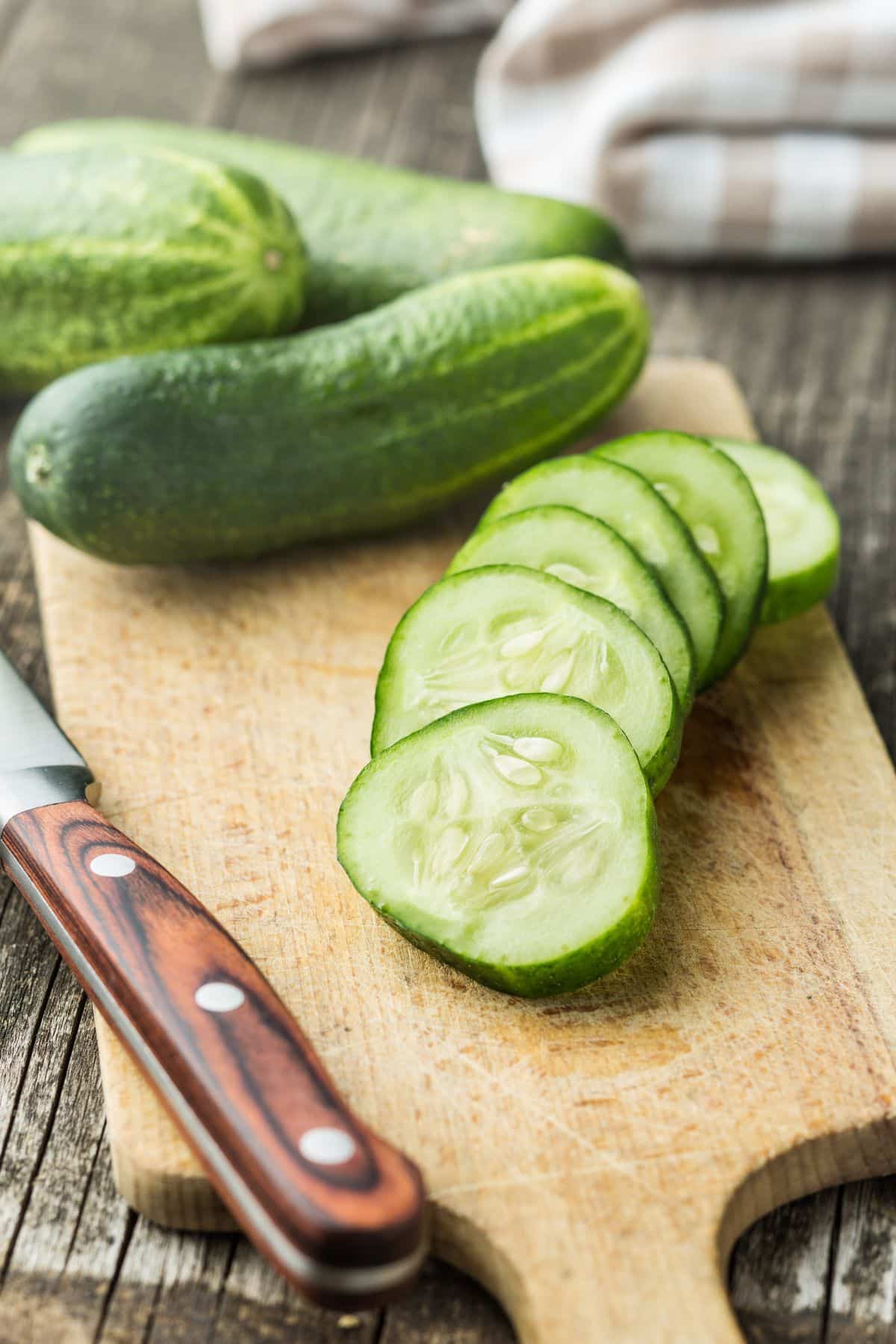 Whole cucumbers and fresh sliced rounds on a wooden cutting board.